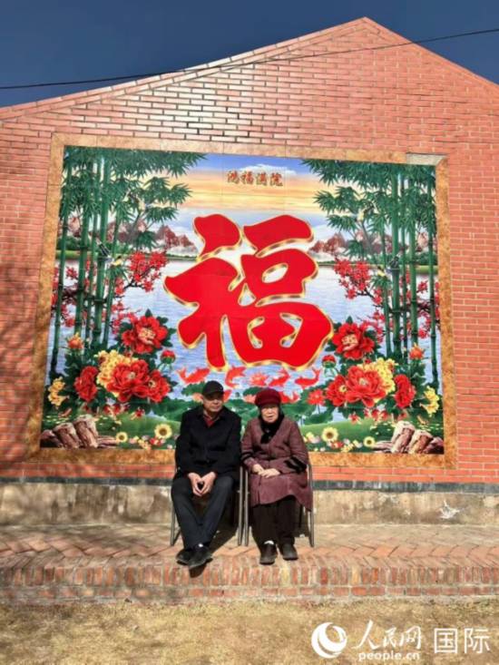 在莫尼山非遗小镇的“福”字墙前，晒着太阳休息的一对老夫妻。人民网 河贞美摄